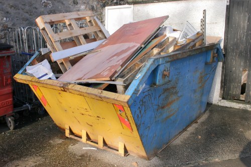A large blue industrial skip situated on a paved area next to a white wall, filled with various construction and renovation waste materials including wooden pallets, painted MDF panels, and metal framing. The skip shows signs of rust and wear on its exterior, with yellow and red reflective safety markings on its sides. The surrounding environment appears to be an outdoor space in Richmond upon Thames, with a damp surface indicating recent rain or cleaning. To the left of the skip, a portion of a metal cart or trolley is visible, and behind it, a black outdoor fence partly encloses the area. The backdrop features a black wooden fence and an adjacent wooden gate. The scene illustrates a typical rubbish removal setup, with Richmond upon Thames House Clearance providing a professional solution for clearing waste debris from residential or commercial properties in the area.