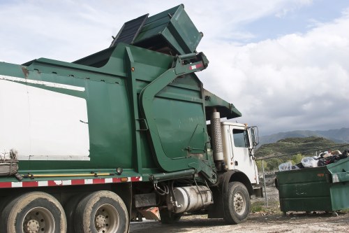A large green and white rubbish collection truck with an open top compactor is parked outdoors on a gravel surface, positioned to the left side of the image. The truck's body is primarily dark green with some lighter green panels and a weathered finish. It features large black tires with visible treads; the rear tires are prominently shown, and the front of the vehicle is partially obscured. The truck's cabin is white with a side mirror and a visible exhaust pipe attached to the side. A green outdoor waste skip filled with mixed rubbish, including black plastic bags and other debris, is situated to the right of the truck, on the gravel. The background reveals a partly cloudy sky over a hilly landscape with low vegetation, indicating a rural or semi-rural setting. The scene captures the process of rubbish removal, consistent with services offered by Richmond upon Thames House Clearance in the local area.